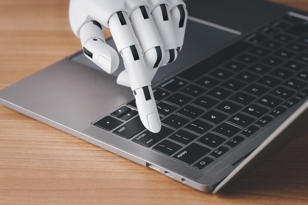 A robotic hand pressing a key on a laptop keyboard while resting on a wooden desk.